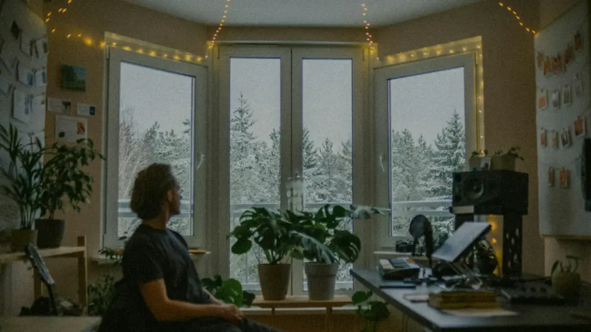 Man looking out window at snowy trees from cozy room.